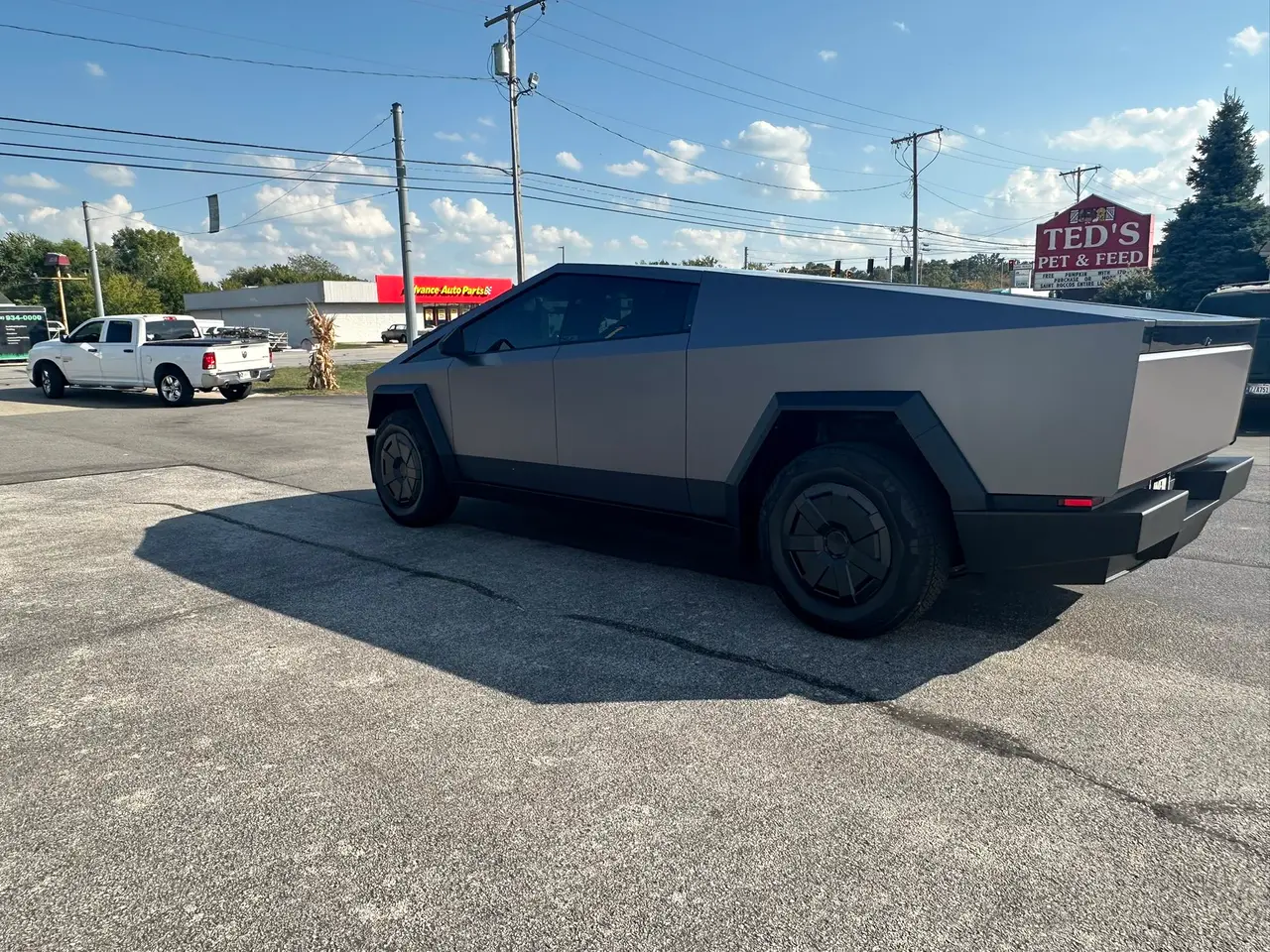 Gray Tesla Cybertruck parked on asphalt on a sunny day. A white pickup truck is in the background.